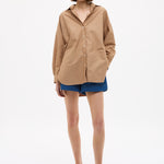 Woman wearing Mara Oversized Long Sleeve Shirt - Toffee with blue shorts and sandals against white background