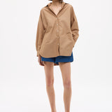 Woman wearing Mara Oversized Long Sleeve Shirt - Toffee with blue shorts and sandals against white background