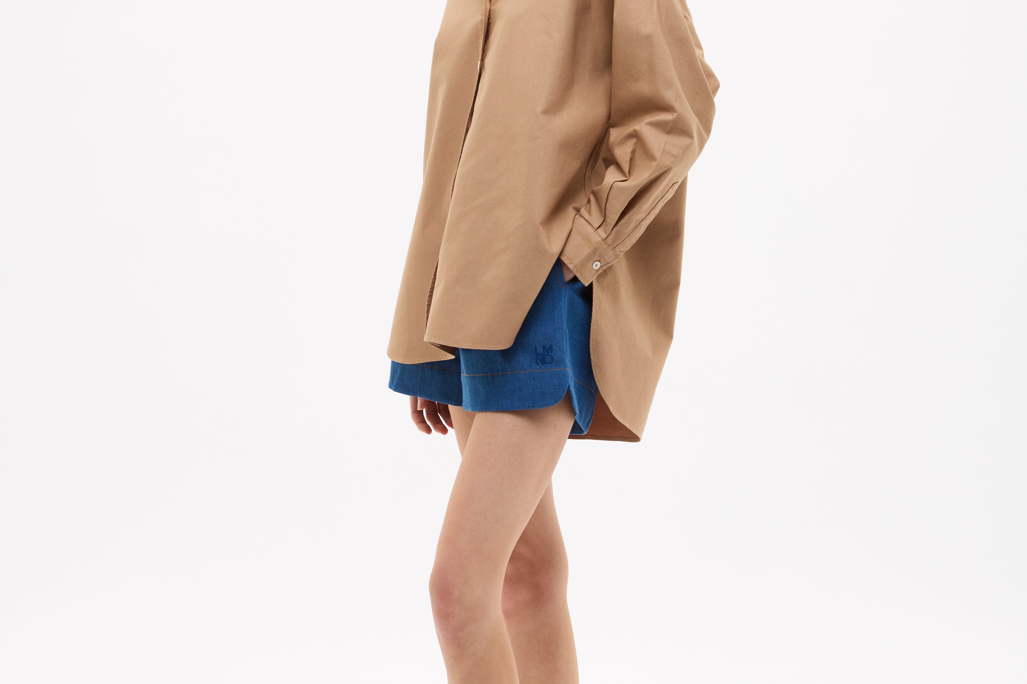 Woman wearing Mara Oversized Long Sleeve Shirt - Toffee with blue shorts and sandals, standing against white background