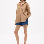 Woman wearing Mara Oversized Long Sleeve Shirt - Toffee with blue shorts and sandals against white background