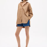 Woman wearing Mara Oversized Long Sleeve Shirt - Toffee with blue shorts and sandals against white background