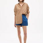 Woman wearing Mara Oversized Long Sleeve Shirt - Toffee with rolled-up sleeves paired with blue shorts and sandals