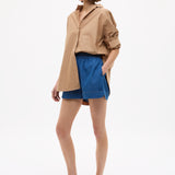 Woman wearing Mara Oversized Long Sleeve Shirt - Toffee with rolled sleeves paired with blue shorts and sandals