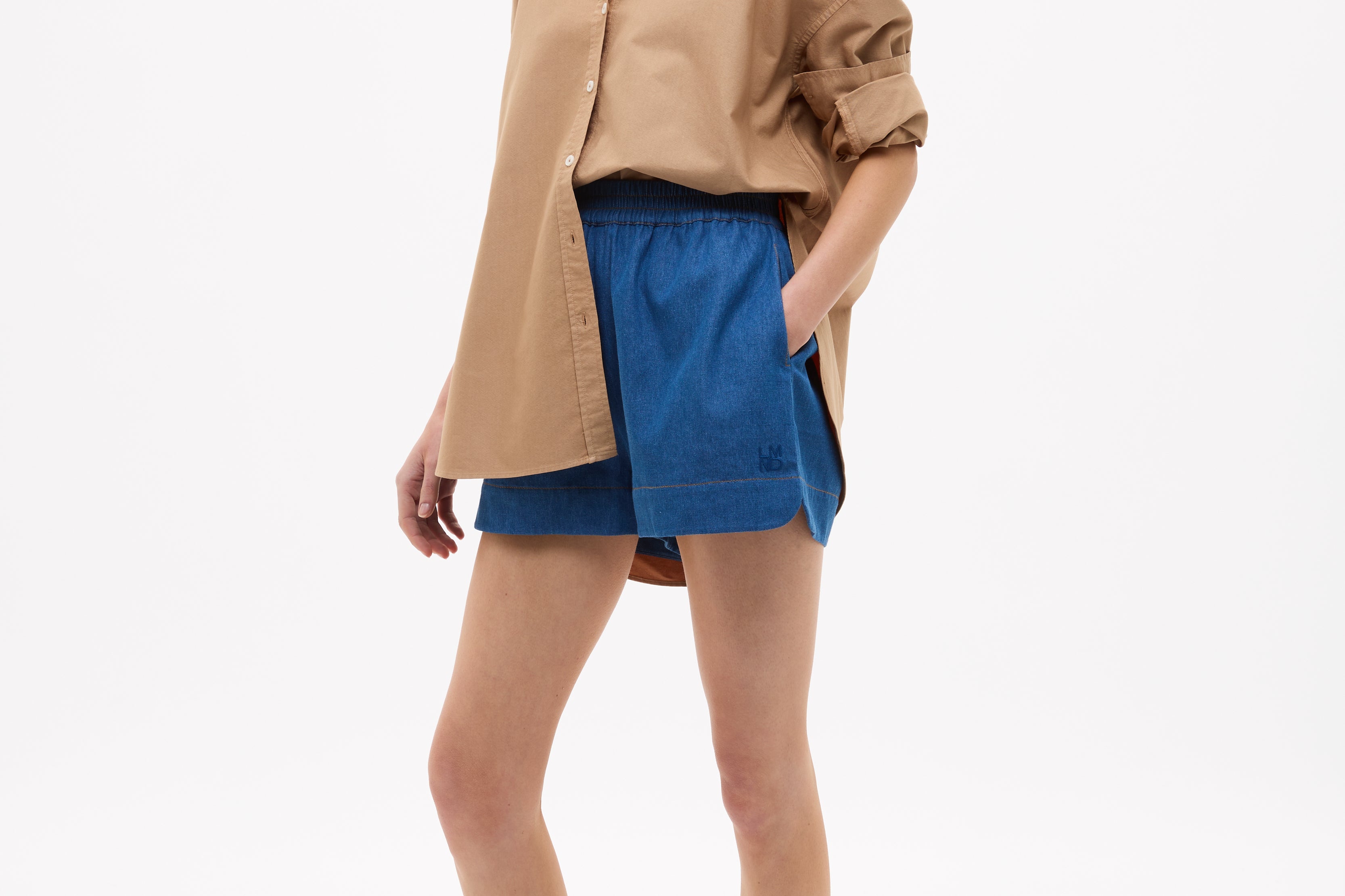 Woman wearing Mara Oversized Long Sleeve Shirt - Toffee with rolled sleeves paired with blue shorts and sandals