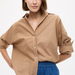 Woman wearing Mara Oversized Long Sleeve Shirt - Toffee with rolled sleeves paired with blue casual pants against white background