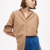 Woman wearing Mara Oversized Long Sleeve Shirt - Toffee tucked into casual blue high-waisted shorts, looking sideways